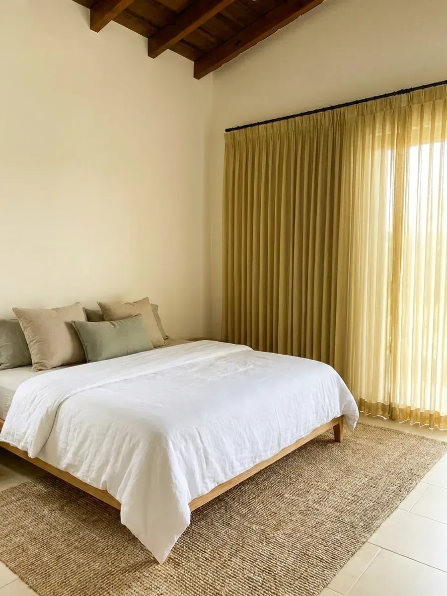 Dormitorio con cortinas de tela sheer color oliva dorado en esquina, cama vestida en lino blanco y alfombra de fibra natural.