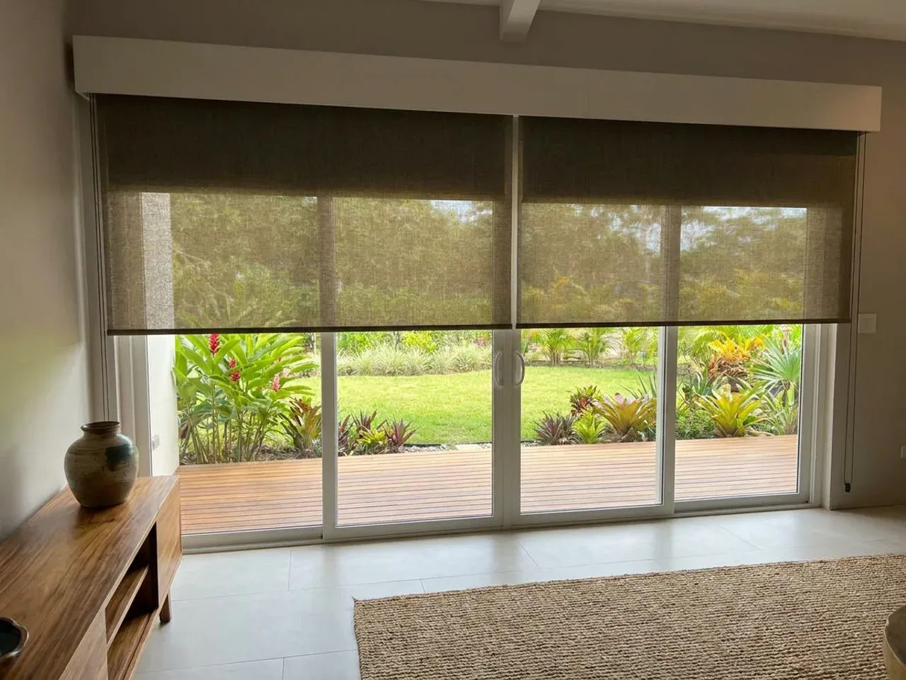 Puerta corrediza con roller Día y Noche gris oscuro bajado a media altura, consola baja de teca y jardín tropical con deck de madera al otro lado.