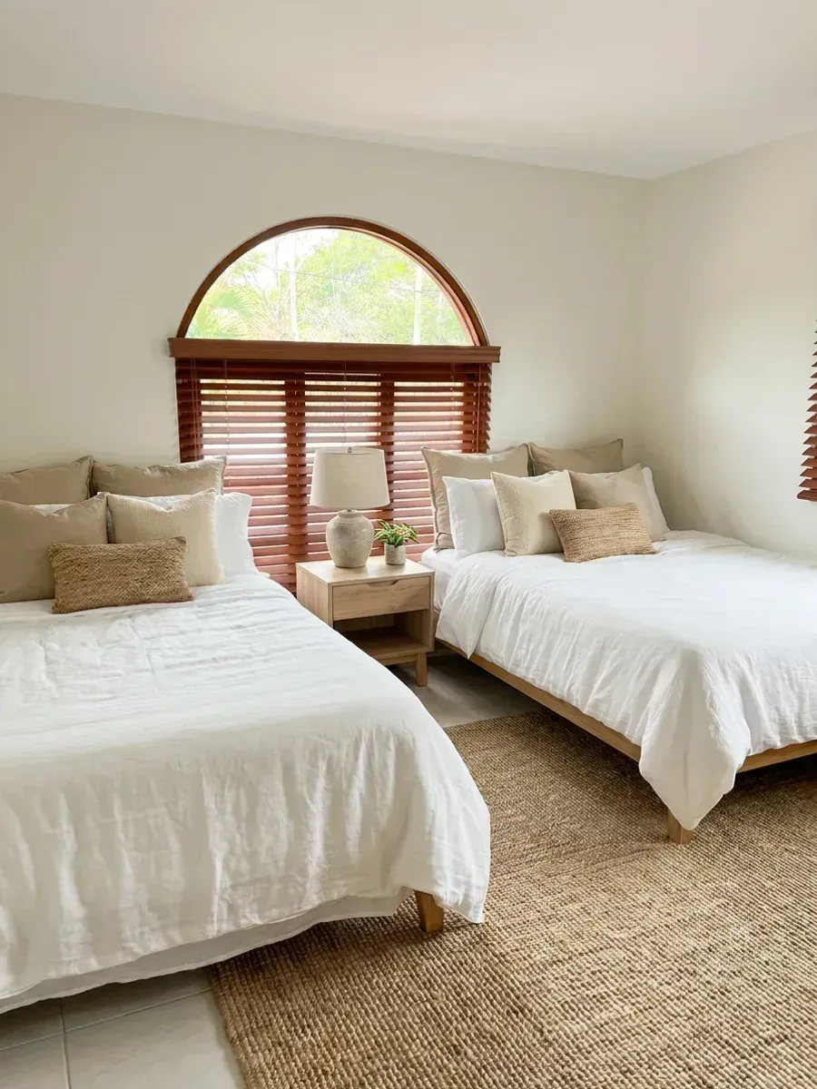 Dormitorio con dos camas y persianas de madera color caoba bajo una ventana de arco, ropa de cama de lino blanco y alfombra de fibra natural.