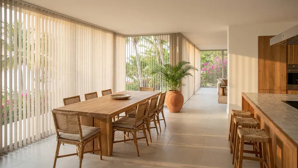 Comedor corrido con persianas verticales de lamas claras a lo largo del ventanal, mesa de teca con sillas de mimbre y barra de cocina de madera.