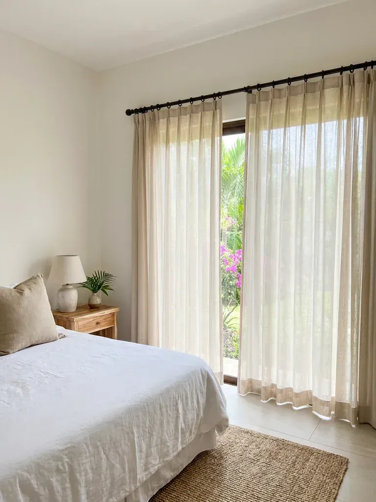 Dormitorio con cortina plisada de lino sobre barral oscuro, cama con ropa de cama blanca, mesa de noche de madera clara y vegetación tropical por el ventanal.