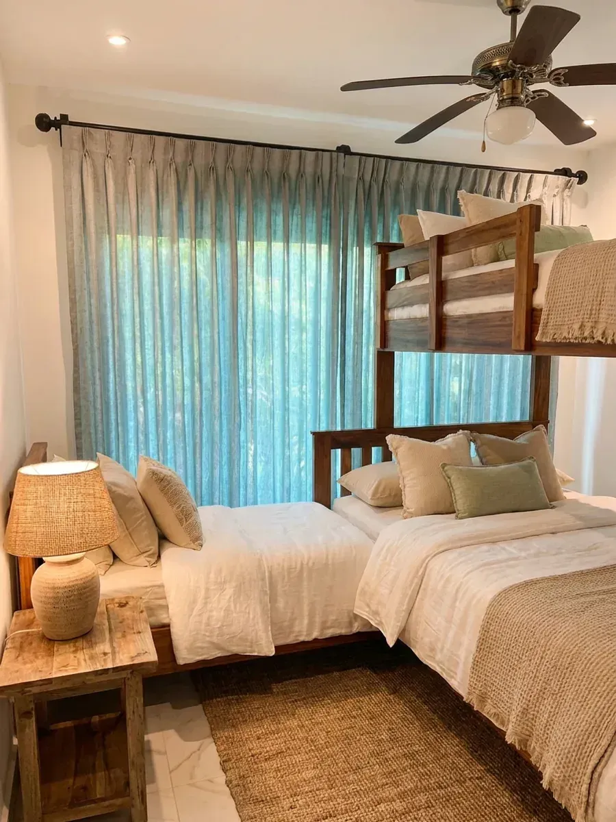 Dormitorio con litera de madera y cortina plisada turquesa corrida sobre la puerta de vidrio, con el verde del jardín insinuándose a través.