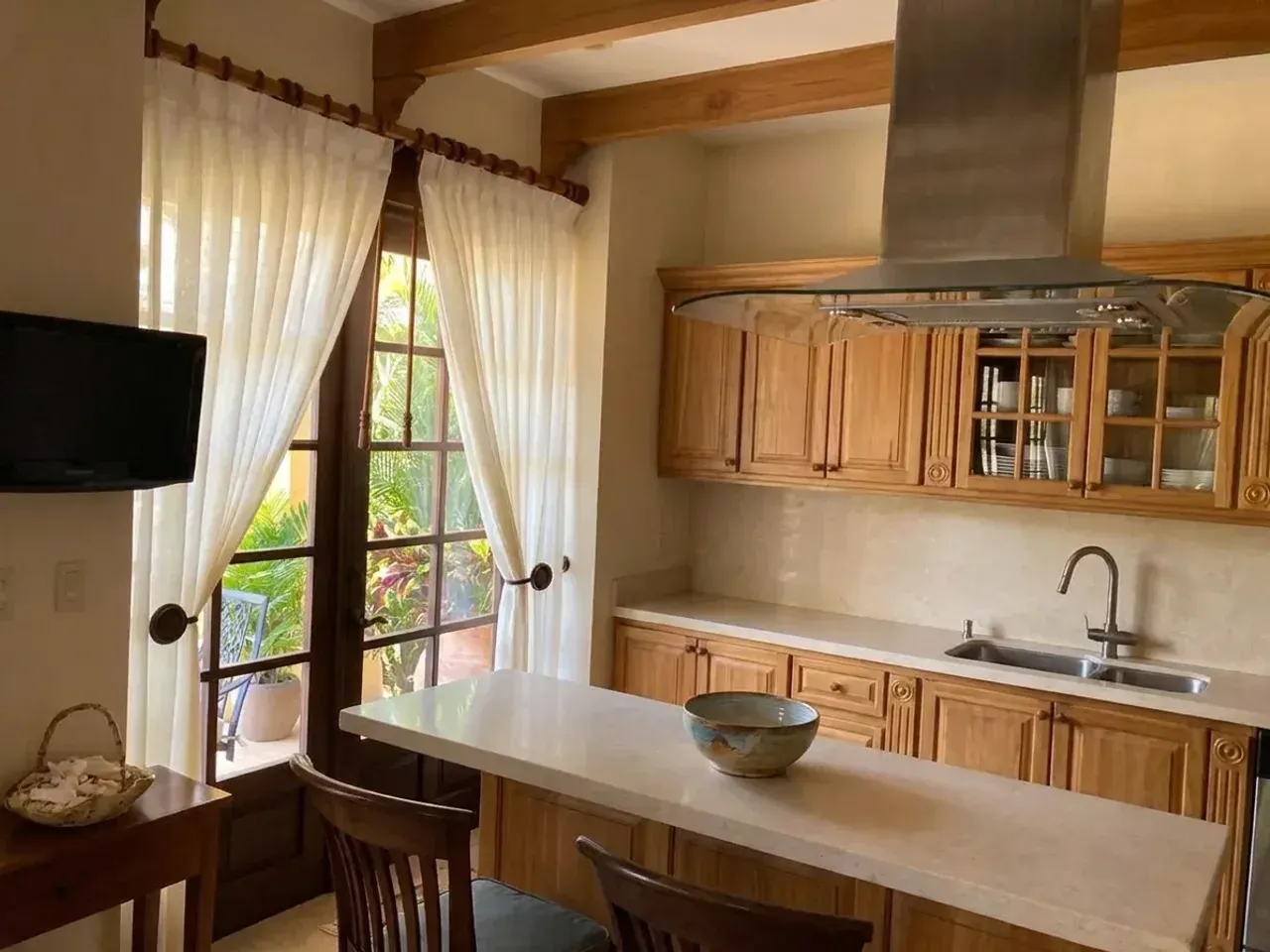 Cocina con muebles de teca clara y encimera de piedra lisa, con cortina plisada blanca recogida a un lado de la puerta de vidrio y vegetación tropical al fondo.