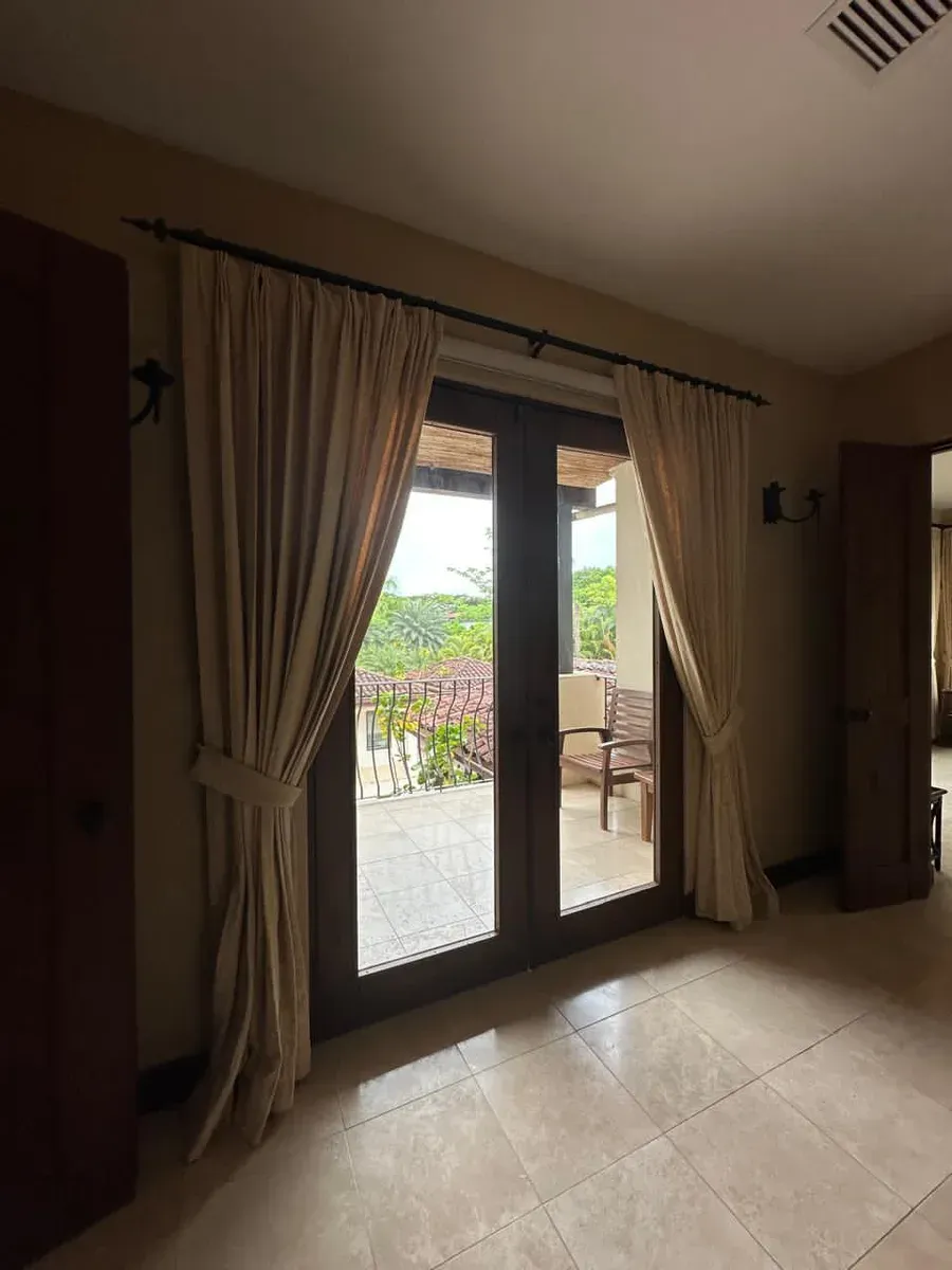 Cortinas plisadas crudo enmarcando puertas francesas oscuras con vista a balcón y vegetación tropical.