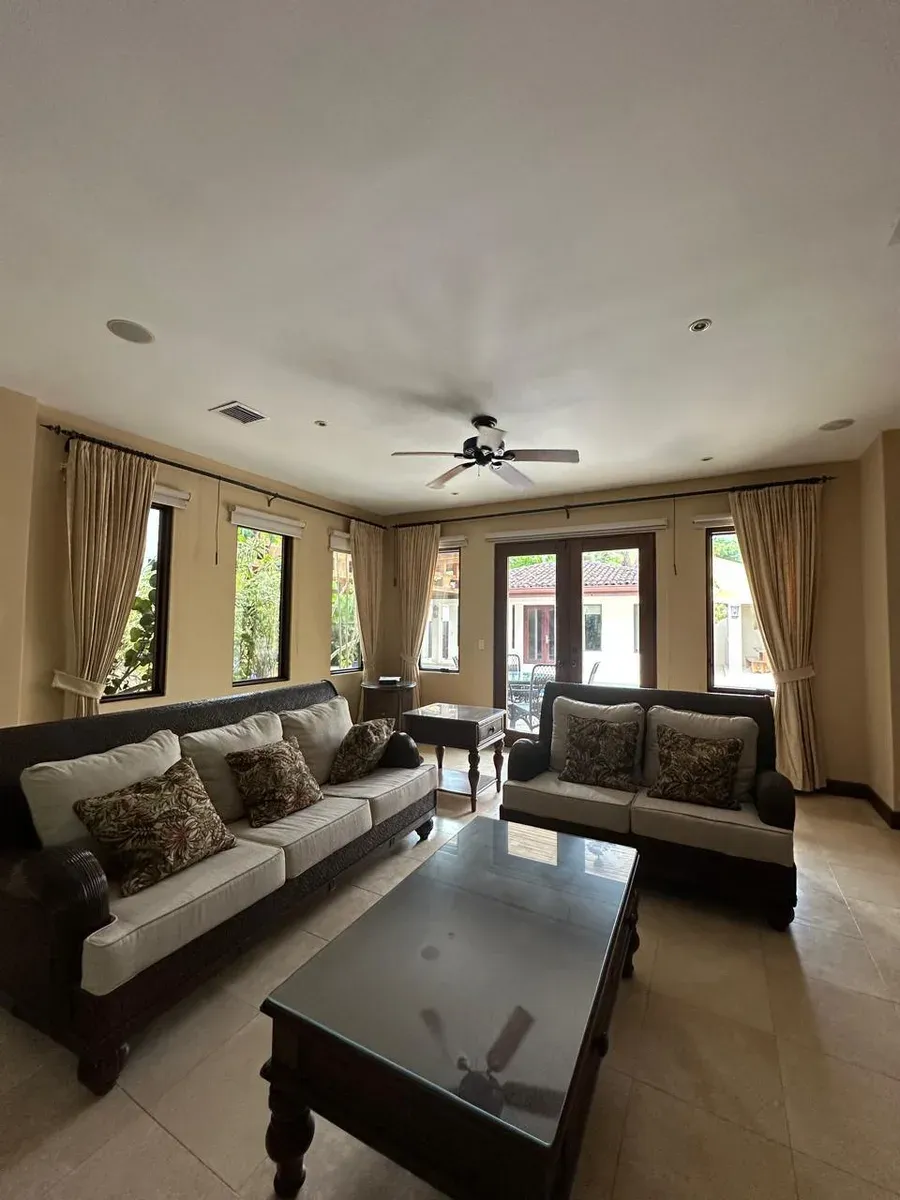 Sala con sofás estampados, ventiladores de techo y cortinas plisadas crema en ventanas y puertas.