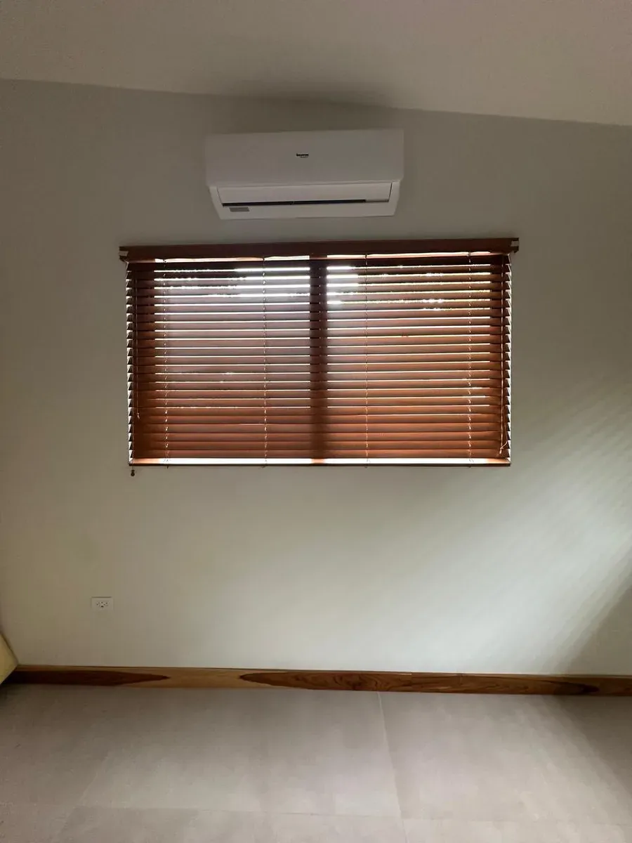 Persianas horizontales de madera en tono caoba sobre ventana, con aire acondicionado split encima.
