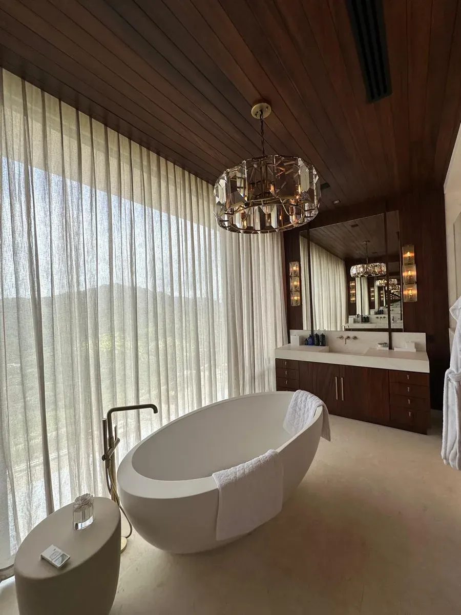Baño con cortinas vaporosas largas en pared completa, tina ovalada y candelabro de cristal facetado.