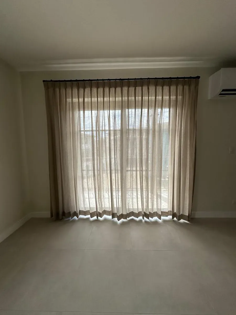 Cortinas plisadas color arena enmarcando ventana amplia, con aire acondicionado split a la derecha.
