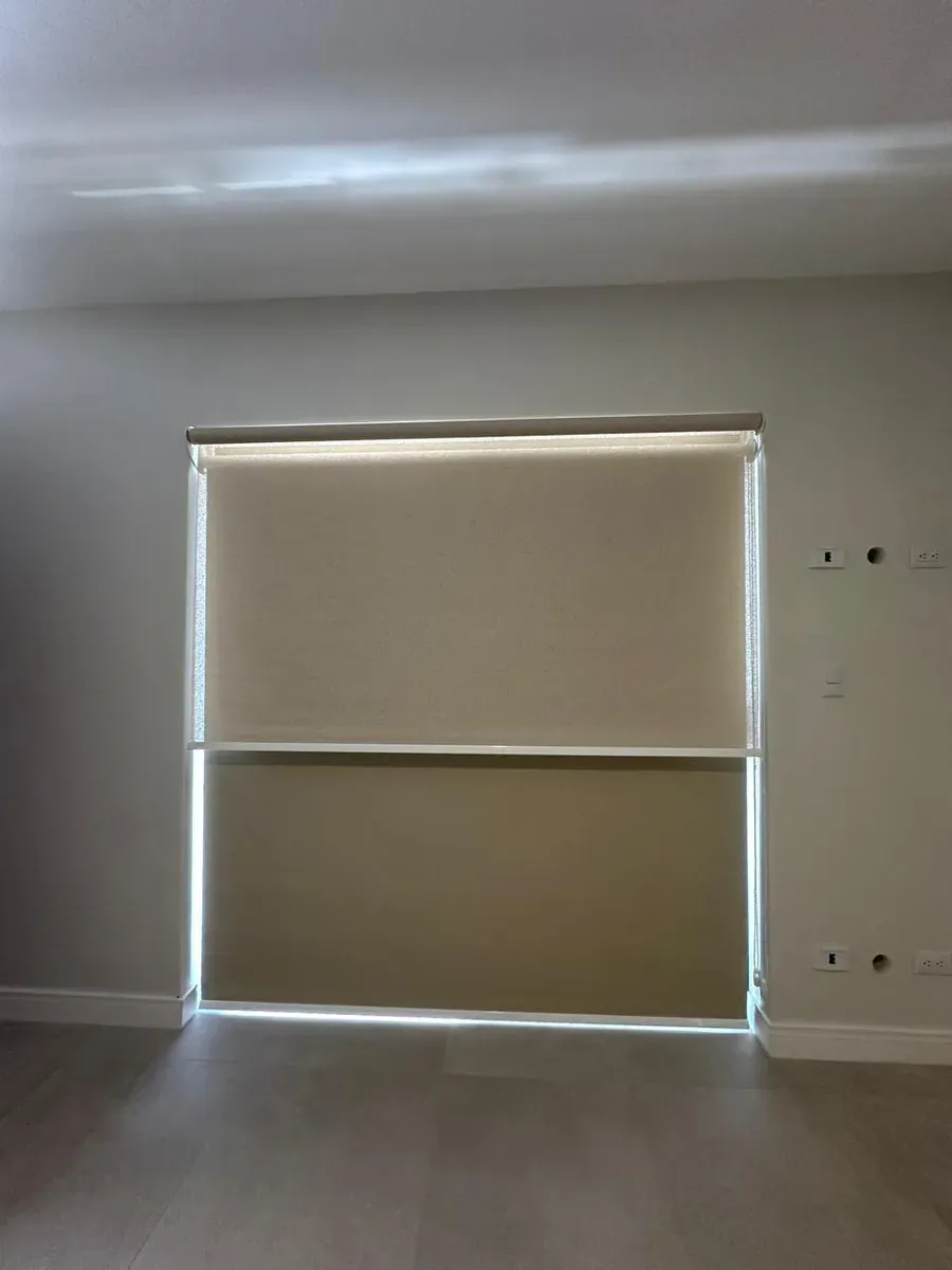 Dos persianas enrollables blackout en tono marfil semi-cerradas con luz filtrándose por los lados.