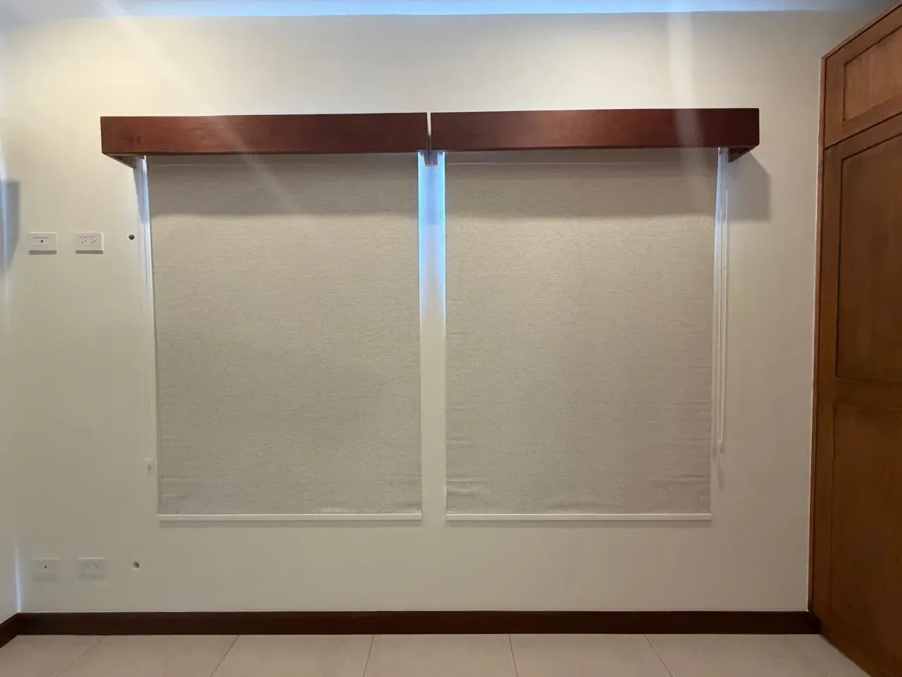Dos persianas enrollables screen crema bajo cornisas de madera oscura sobre pared blanca.