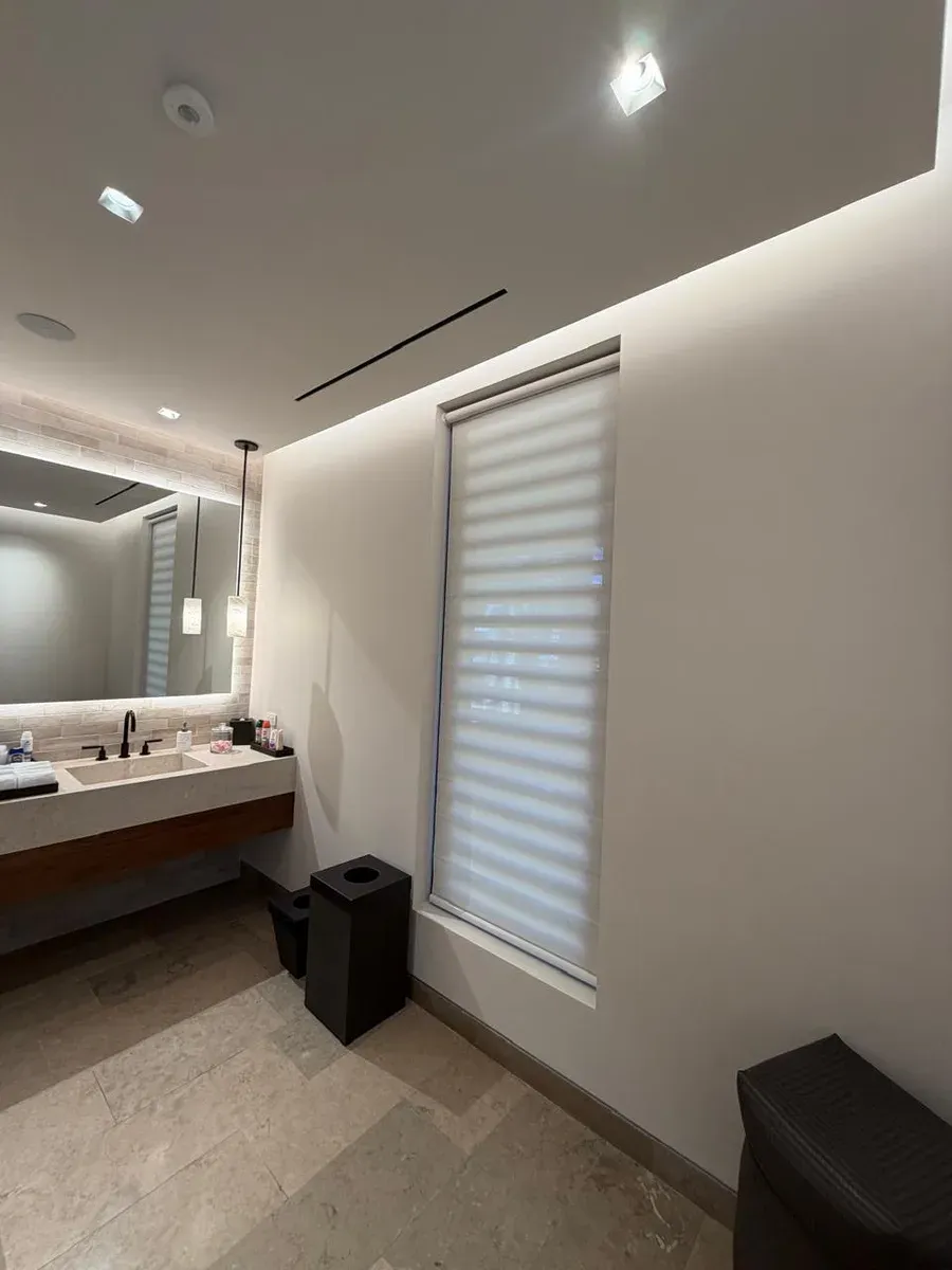 Baño moderno con persiana día y noche blanca, lavabo de concreto y espejo con luz indirecta.
