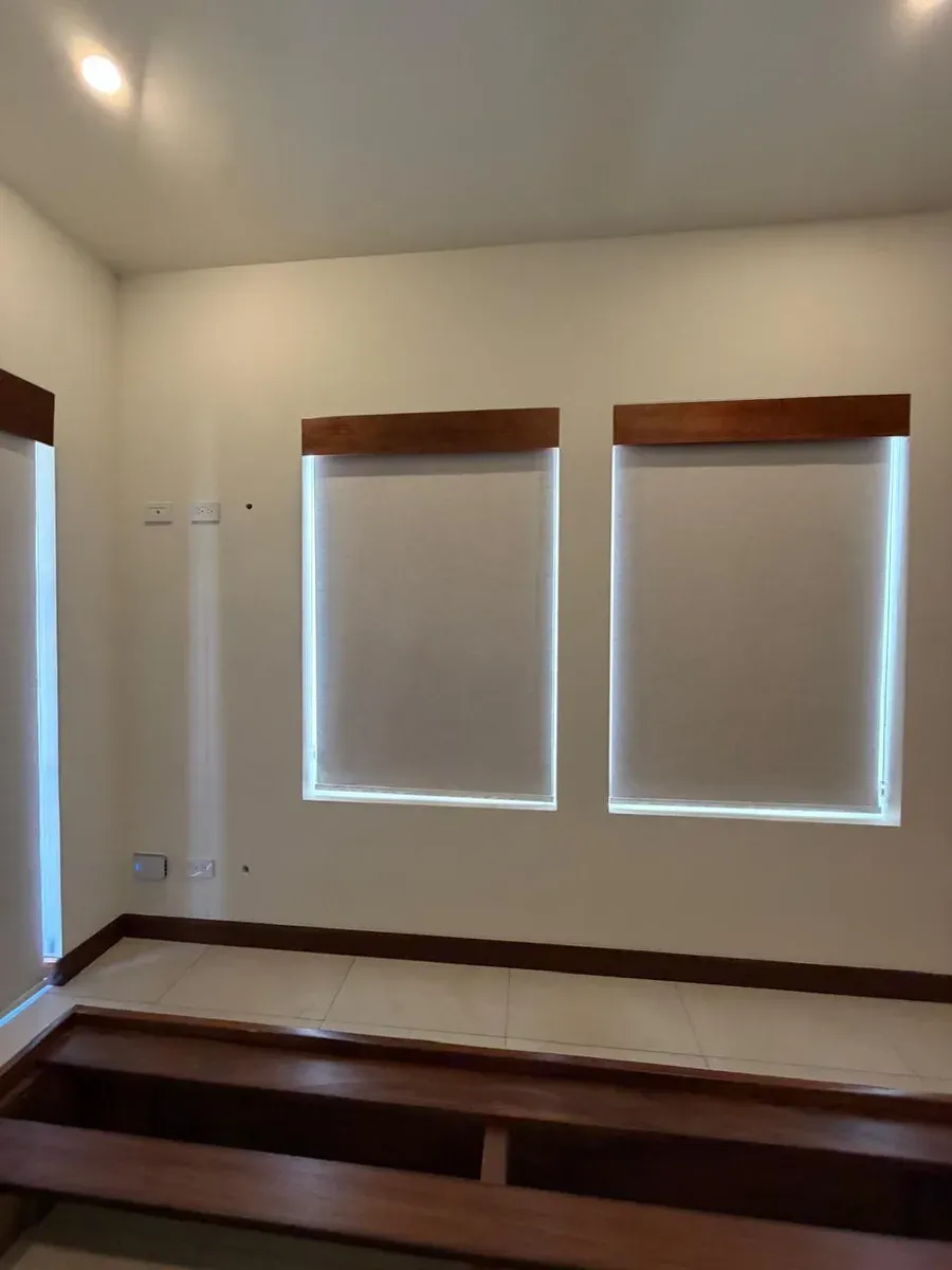 Dos persianas enrollables screen crema con cornisas de madera oscura sobre ventanas pareadas.