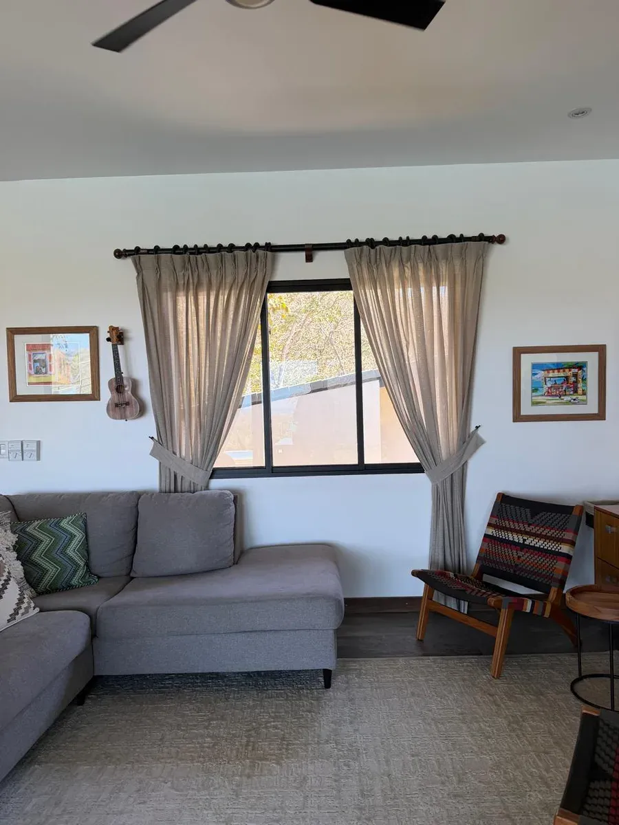 Sala con sofá gris seccional, cortinas plisadas arena con alzapaños y vista a árboles tropicales.