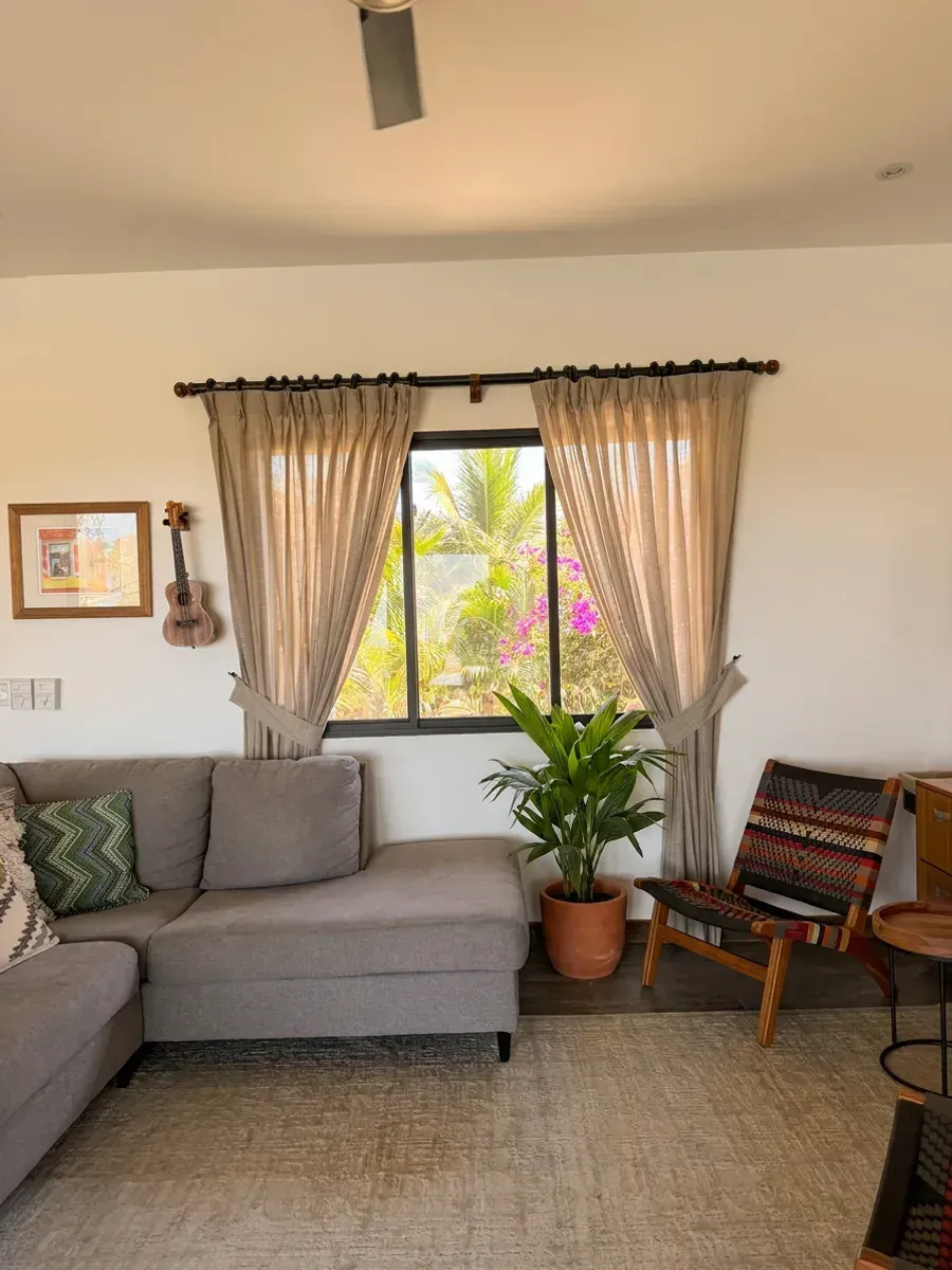 Sala con sofá gris seccional, cortinas plisadas arena con alzapaños y vista a árboles tropicales.