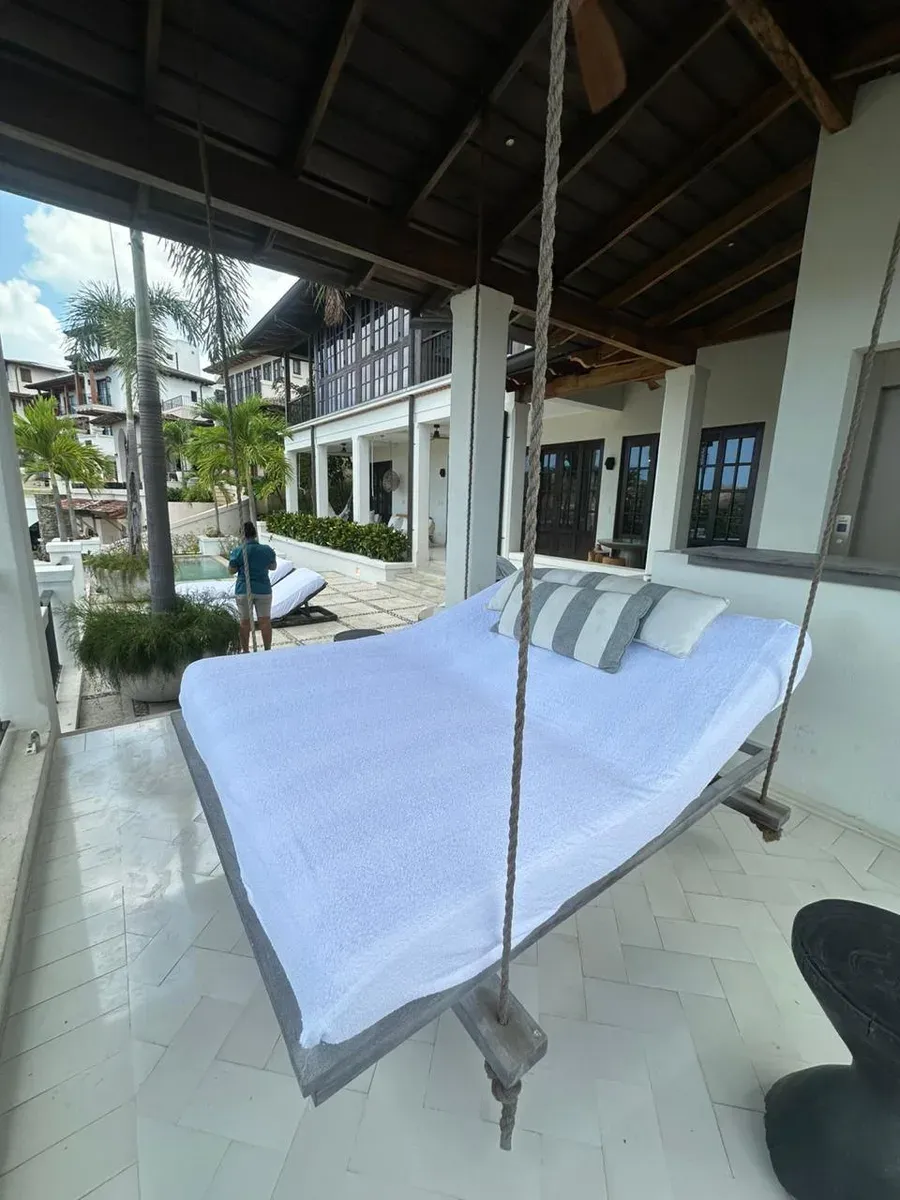 Terraza cubierta con cama colgante de cuerdas trenzadas, cojines rayados y vista a vecindario colonial.