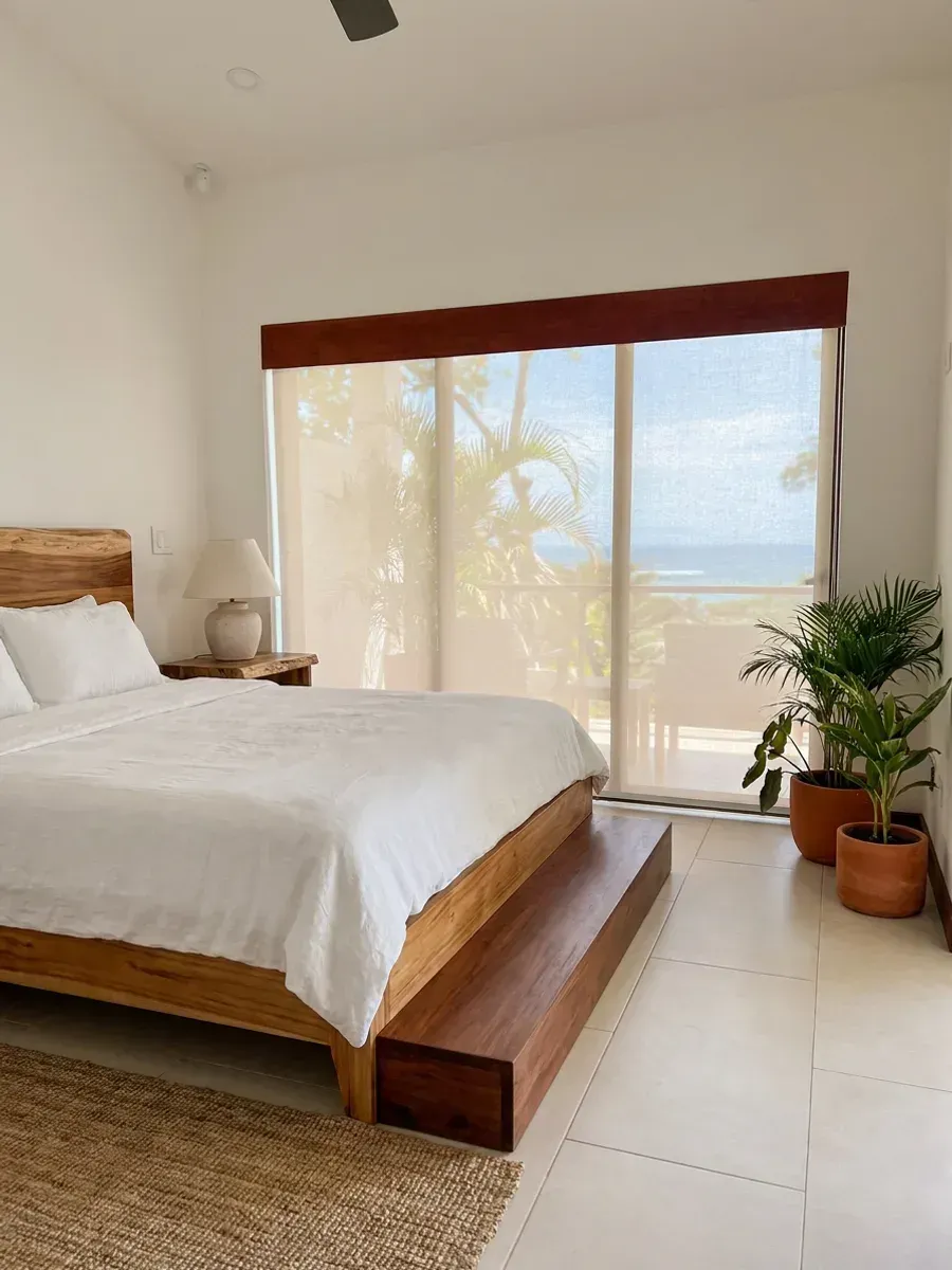 Dormitorio con persiana enrollable screen crema bajo cornisa de madera oscura, cama de madera vestida en lino y el escalón de tablones a los pies.