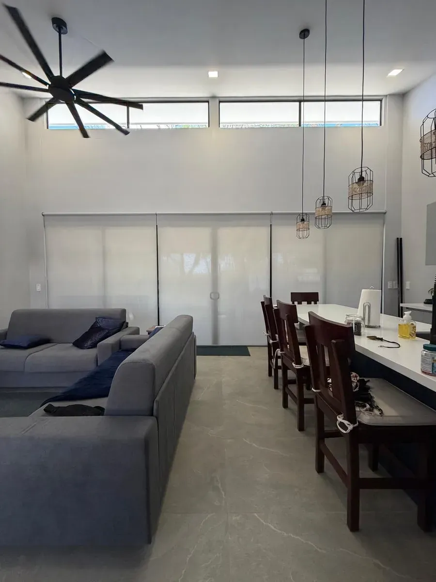 Sala open-plan con persianas screen a doble altura, ventilador industrial y barra blanca con sillas oscuras.