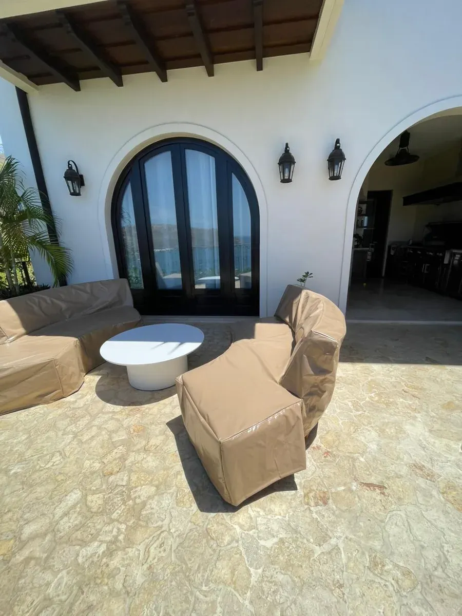 Patio colonial con muebles enfundados, mesa redonda blanca y puerta francesa en arco con vista al mar.