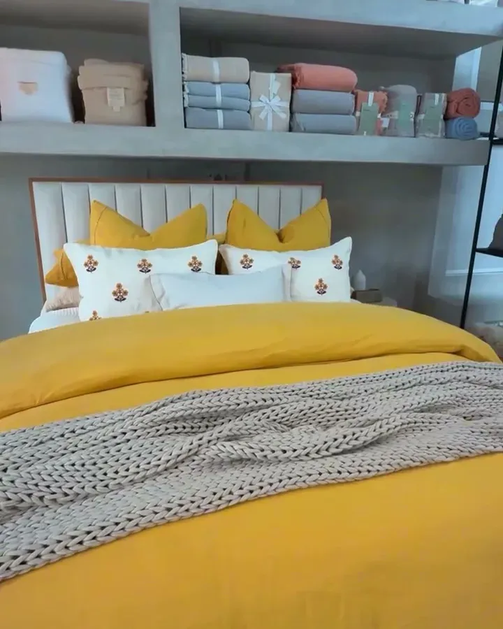 Bed dressed with a yellow duvet, cable-knit throw, and embroidered pillows, in front of the bedding shelves in the showroom.