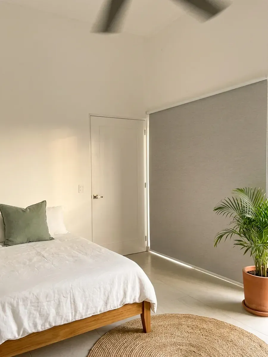 Cuarto de niños con Sun Screen cubriendo el vano entero, cama baja de madera clara con ropa de cama de lino, alfombra redonda de fibra natural y planta en maceta de barro.
