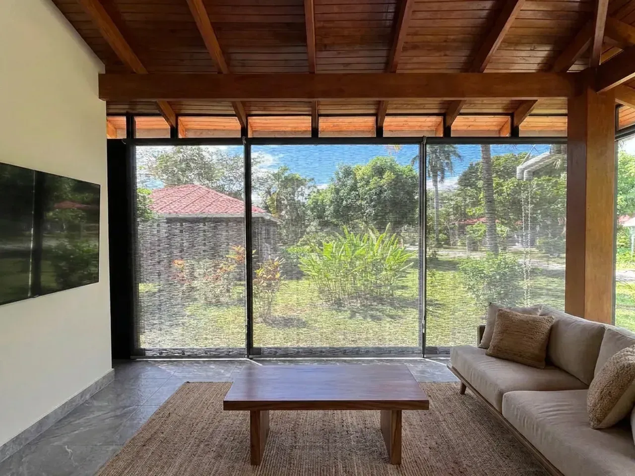 Sala bajo techo de vigas de madera con Sun Screen sobre el ventanal corrido, sofá de lino arena, mesa de centro de madera maciza y jardín visible a través del tejido.