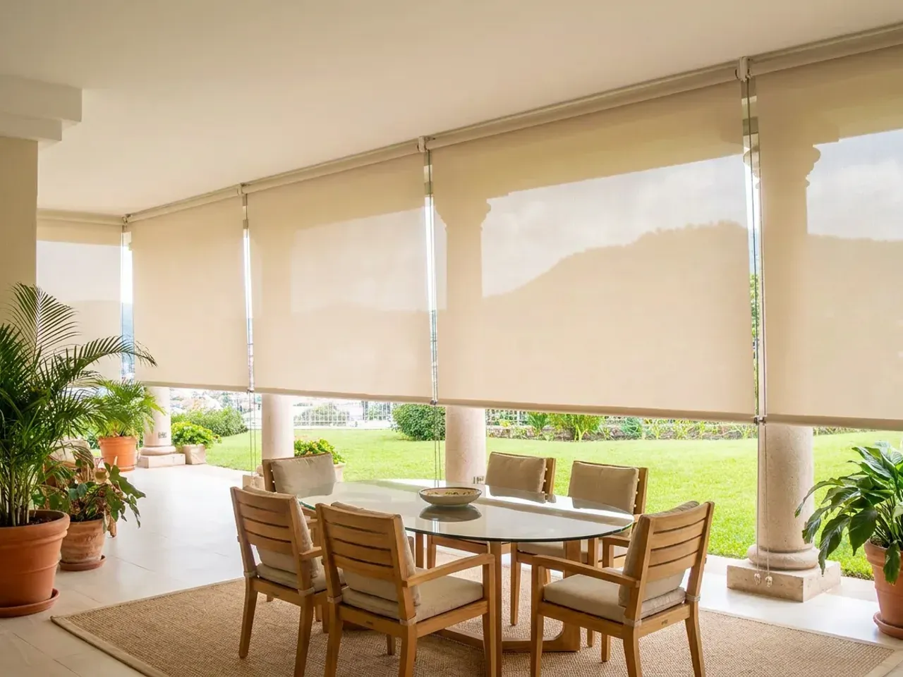 Terraza techada con columnas y una corrida de cortinas de toldo bajadas hacia el jardín, mesa redonda de teca con sillas y alfombra de fibra natural.