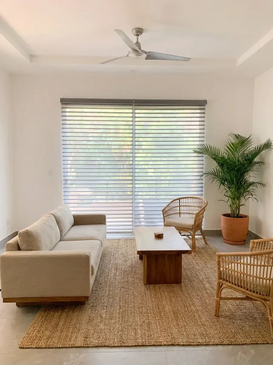 Sala luminosa con Triple Shade de láminas horizontales sobre el ventanal, sofá de lino y palmera en maceta de barro.