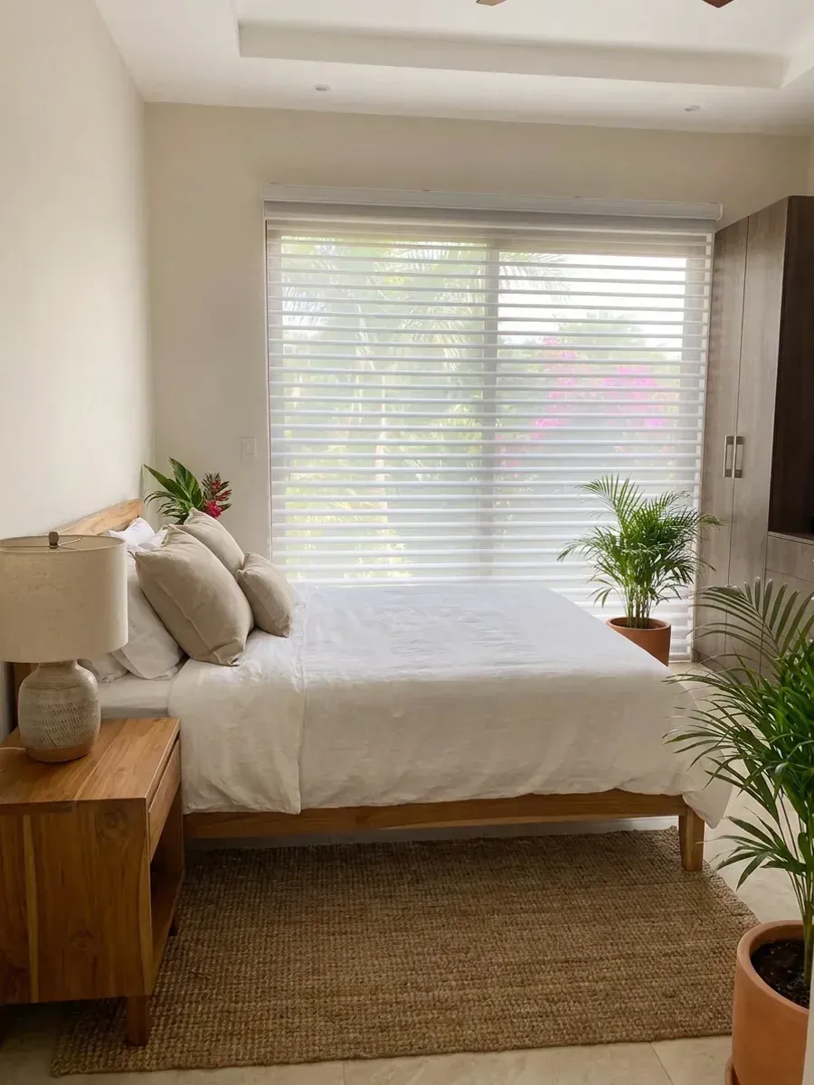 Dormitorio con Triple Shade sobre el ventanal, cama de madera clara con ropa de cama blanca, alfombra de fibra natural y plantas en maceta de barro.