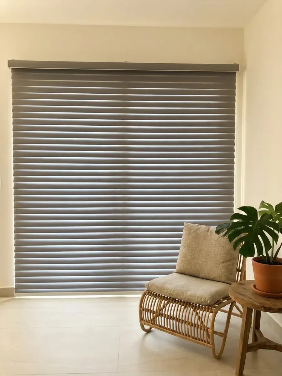 Triple Shade gris cerrado sobre el ventanal, con butaca de fibra natural, mesa auxiliar de teca y planta en maceta de barro.